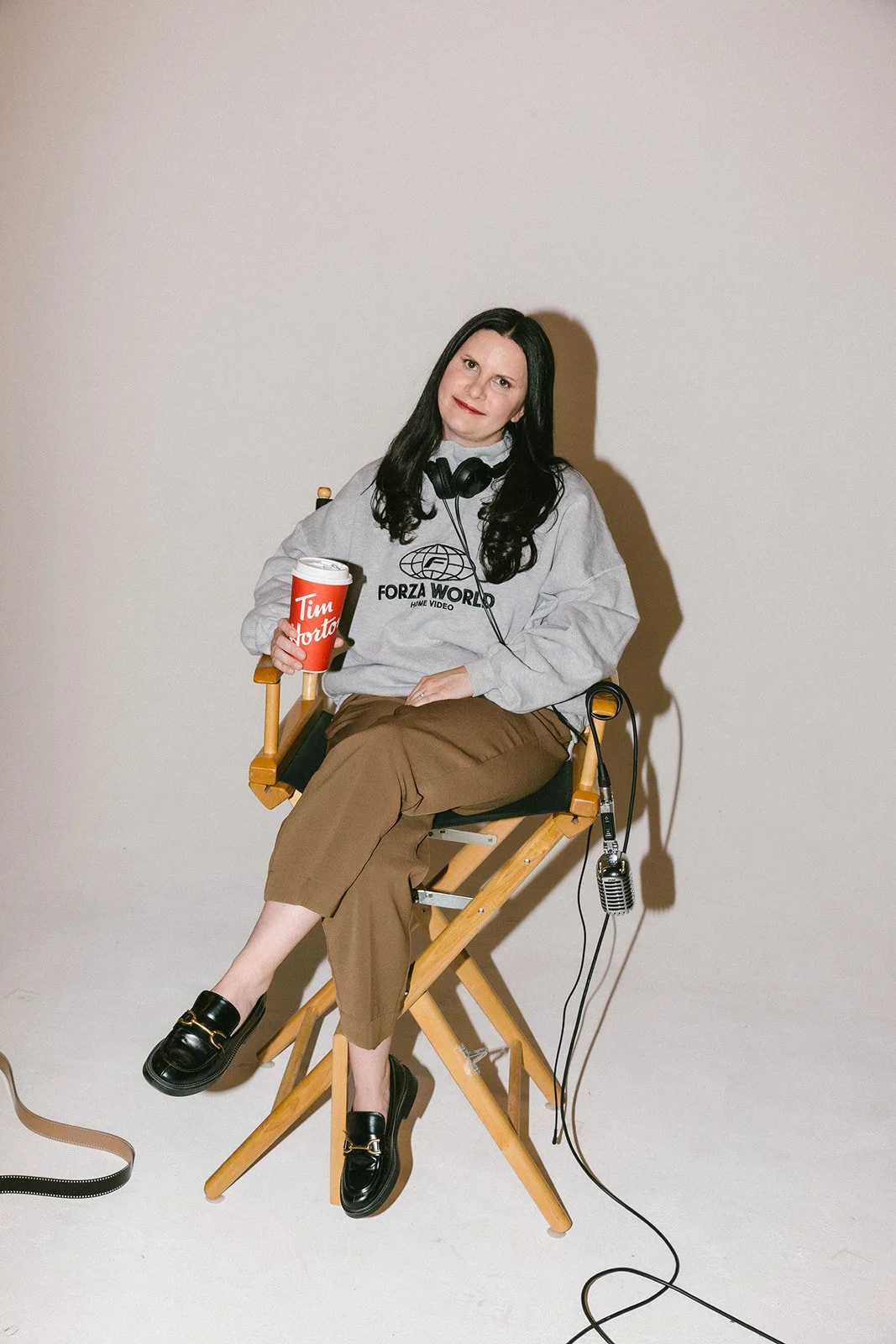 A woman with black hair sitting on a director's chair, holding a Tim Hortons cup, wearing a gray hoodie with 'Forza World' logo, brown pants, and black loafers, with headphones around her neck and a microphone on the floor.