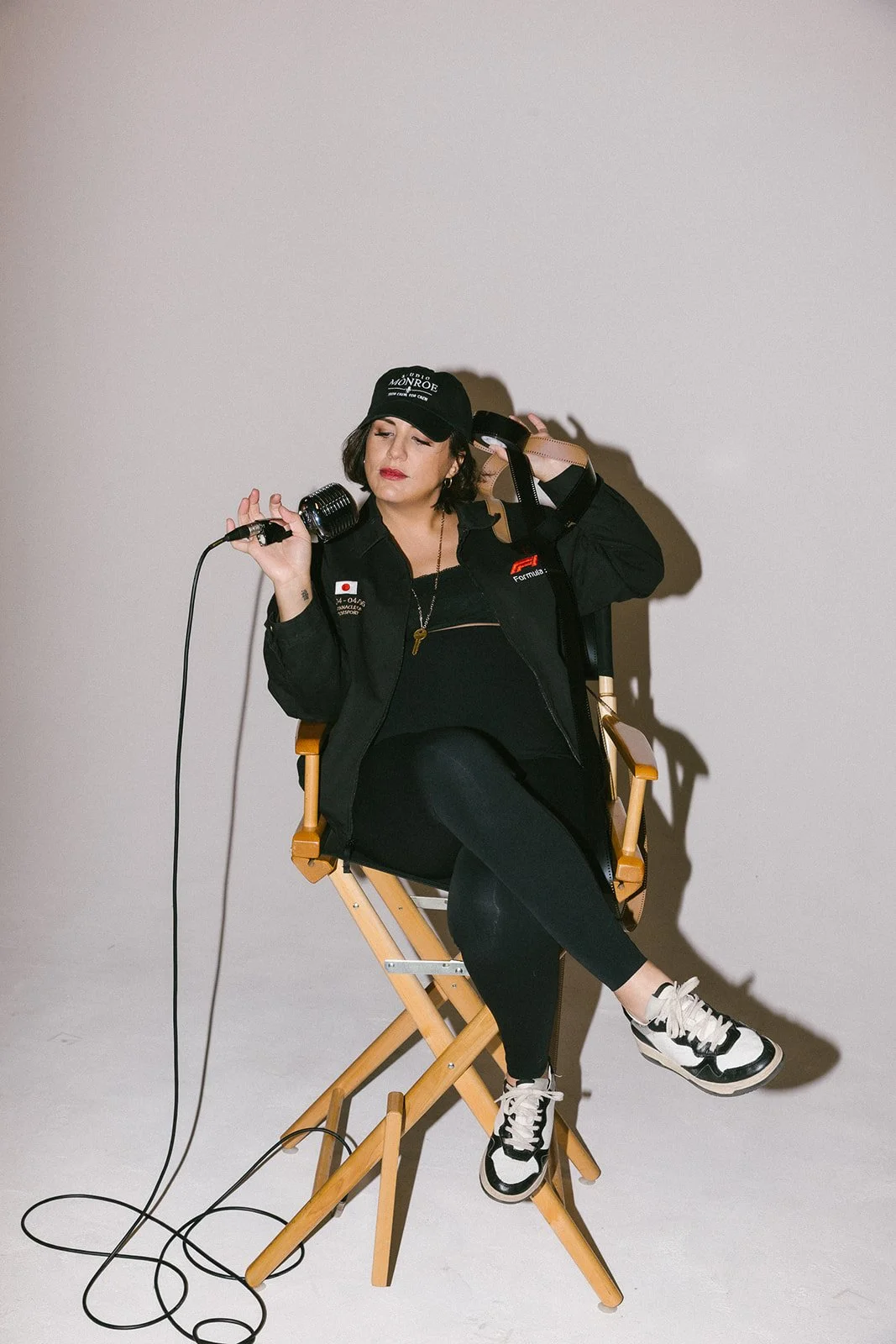Woman sitting on a wooden chair, holding a vintage microphone, wearing a black cap, black jacket, black pants, and sneakers, with a plain white background.