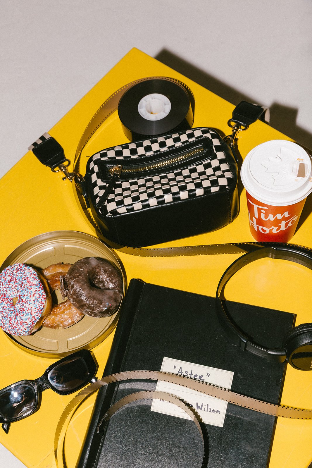 A yellow table with various items including a black and white checkered handbag, a Tim Hortons cup, a plate of donuts, a book titled "Aster" by Wilson, a pair of sunglasses, a black headphone, and a roll of black tape.