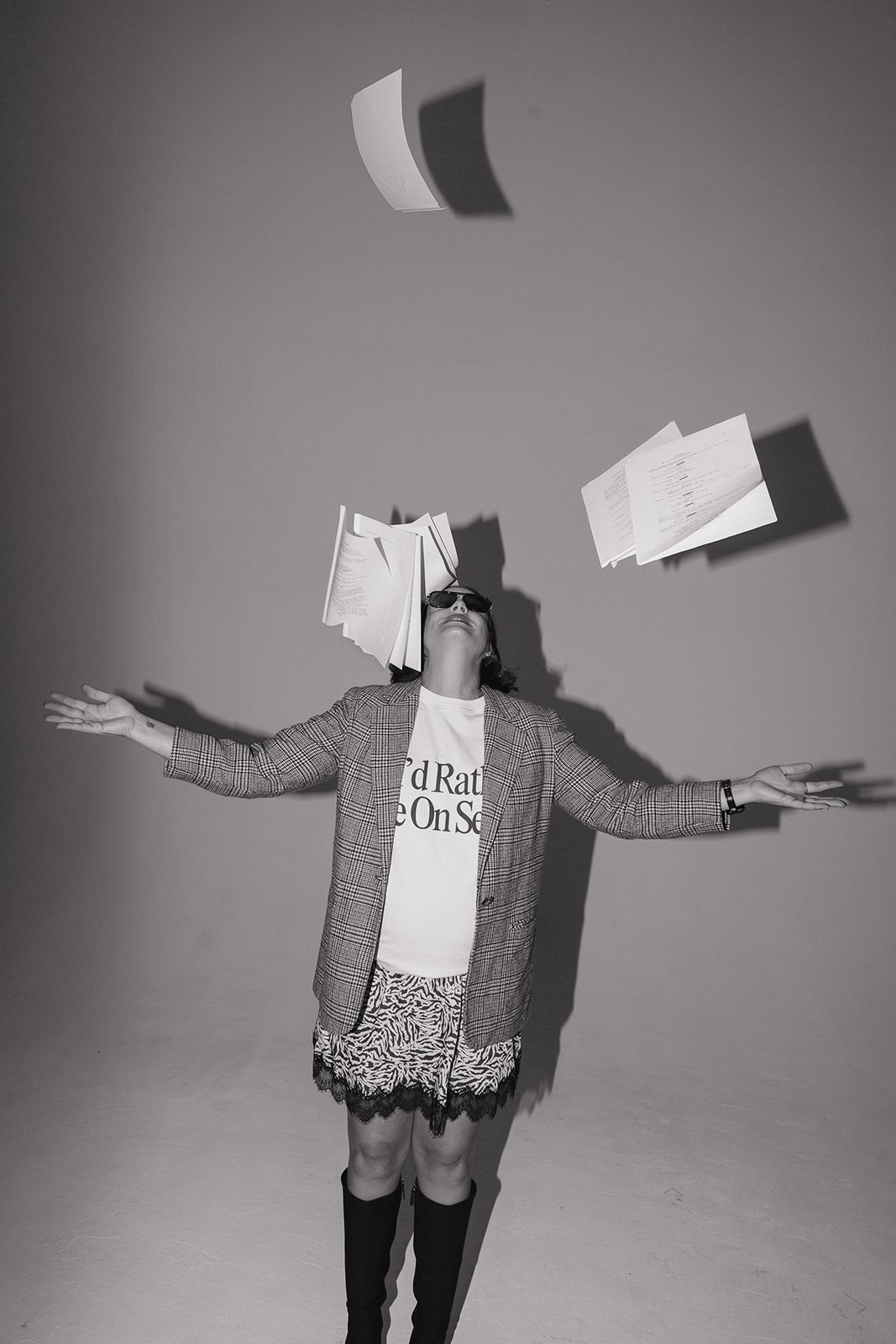 Person wearing sunglasses with papers flying over head, arms outstretched, wearing a patterned blazer, skirt, and boots, in a studio with plain background.