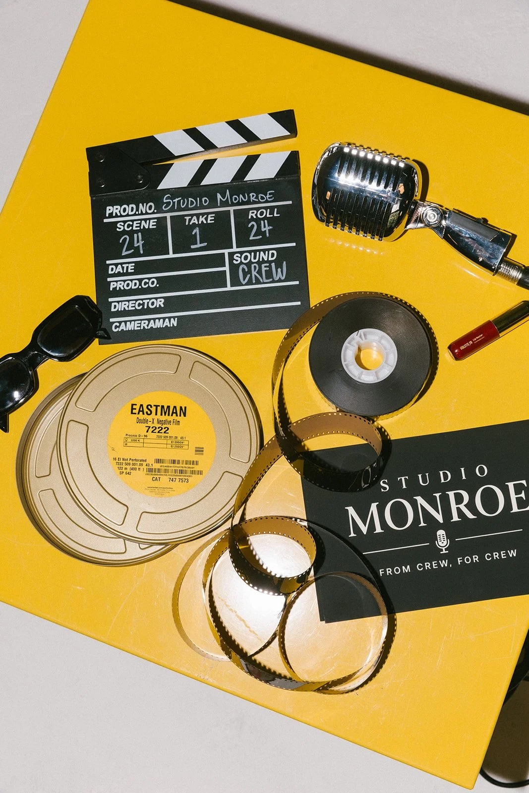 Film reel, clapperboard, vintage microphone, film strip, sunglasses, tube of lipstick, and a yellow background with a Studio Monroe booklet.
