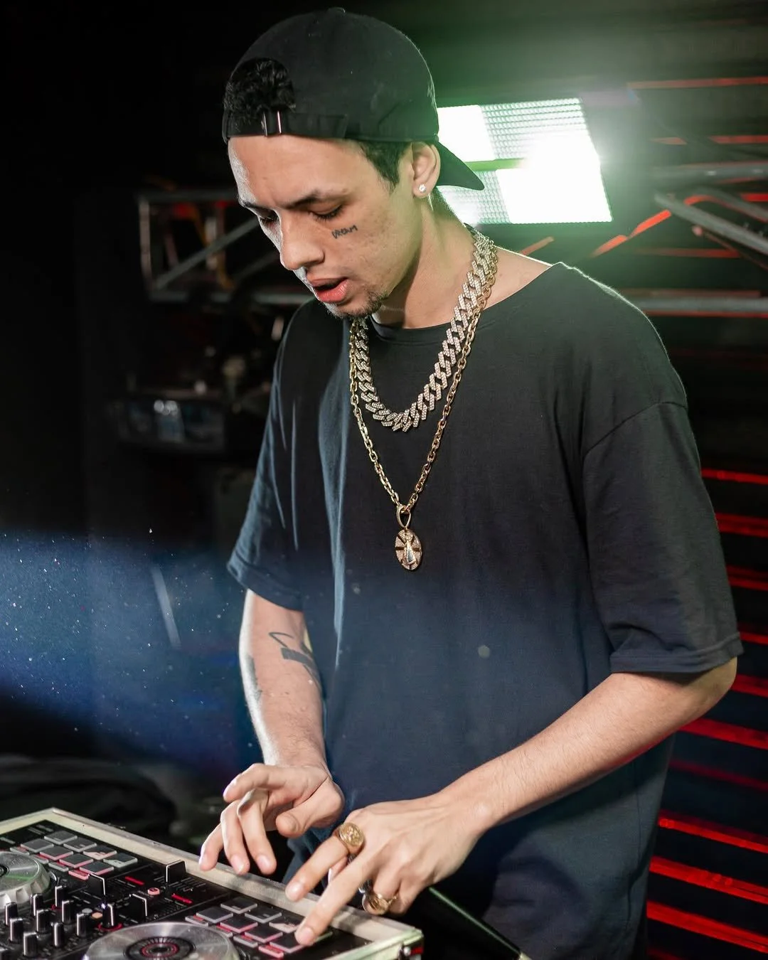 Homem de casaco preto e boné preto, com várias correntes douradas, usando brincos e anéis, operando um equipamento de DJ em um ambiente com luzes brilhantes.