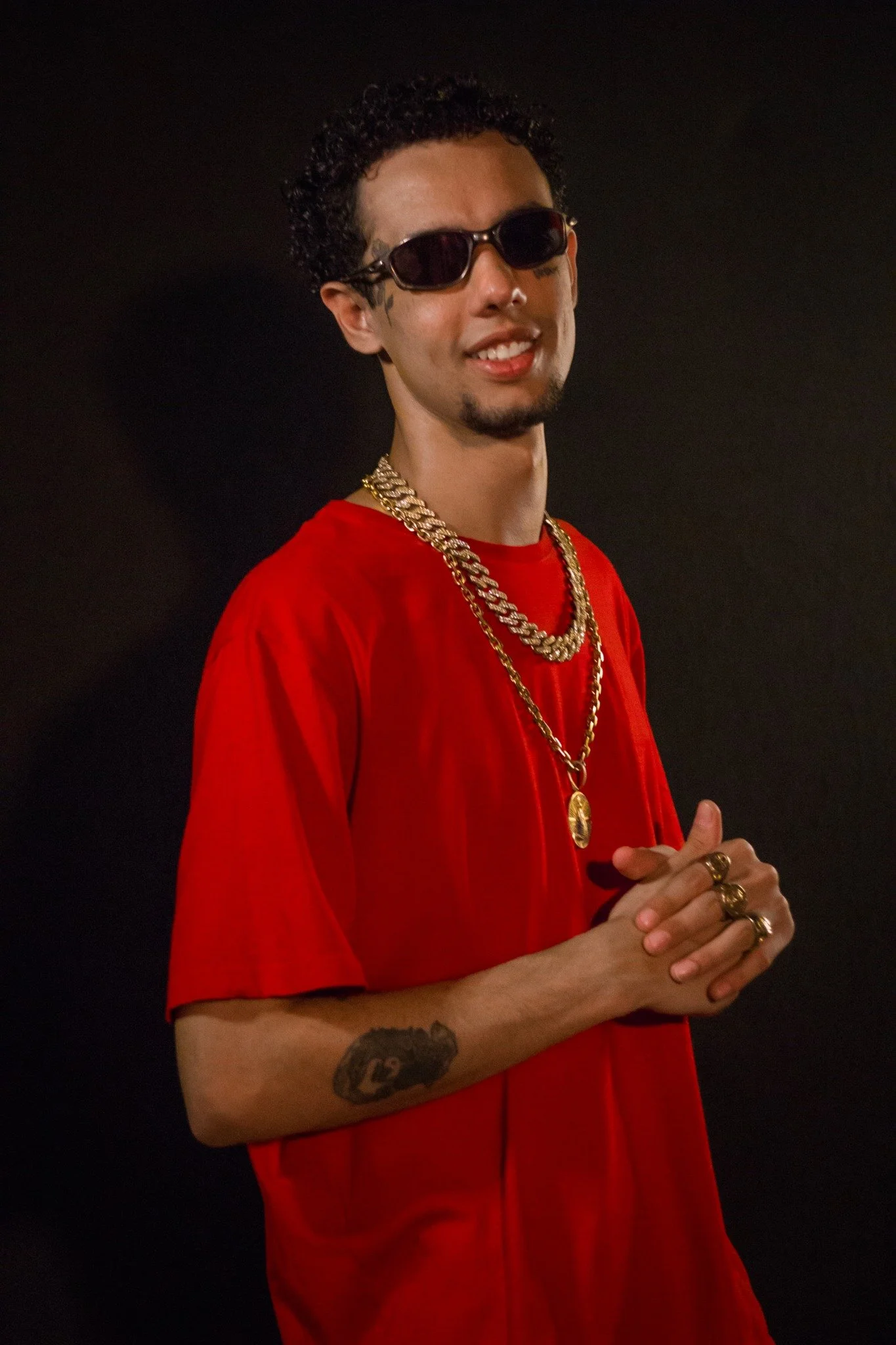 Homem com cabelo cacheado, usando óculos escuros, camiseta vermelha, várias correntes douradas, anéis dourados e tatuagem no braço, posando contra fundo preto.