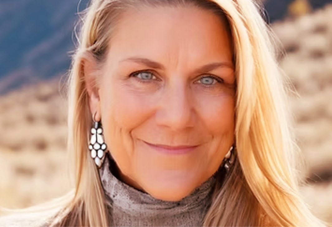 Close-up of a woman with blonde hair and blue eyes, smiling, wearing earrings and a turtleneck outdoors with a blurred desert landscape in the background.