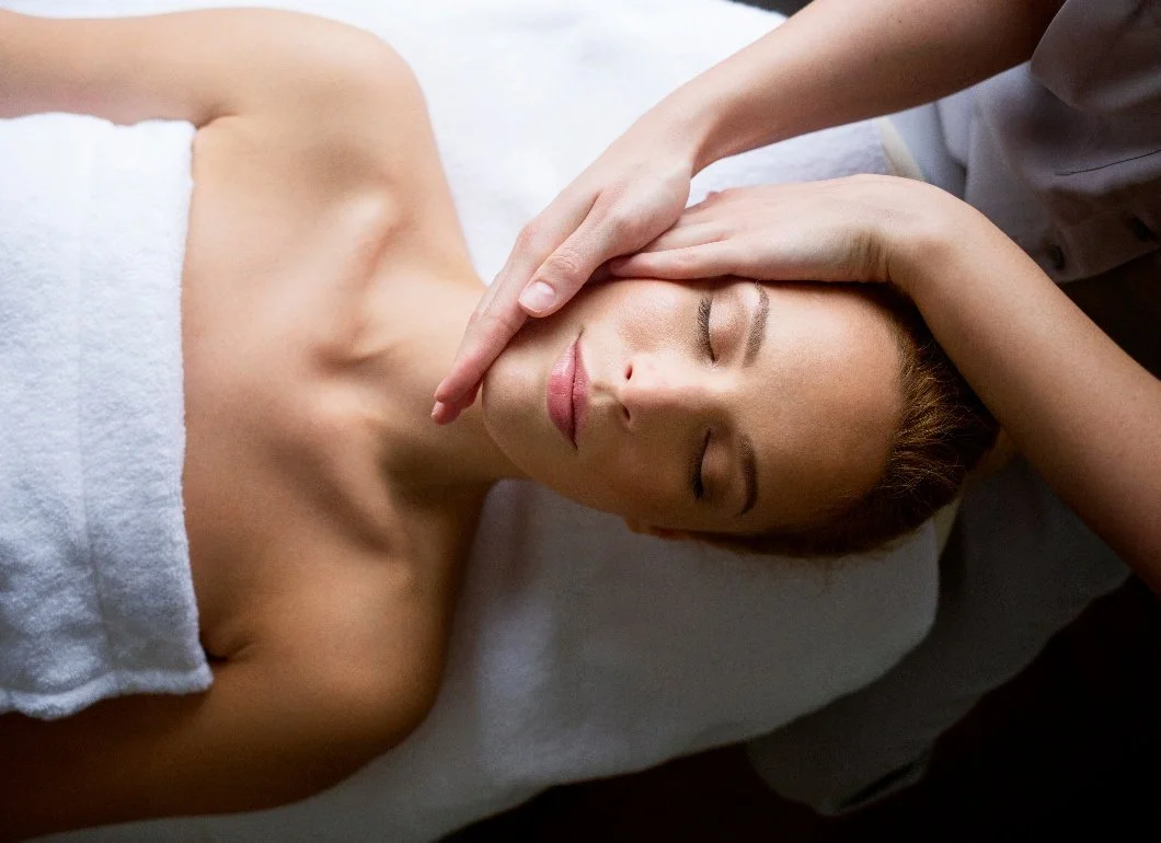 A woman receiving a facial massage at a spa or massage therapy session, lying on her back with her eyes closed, covered with a towel.