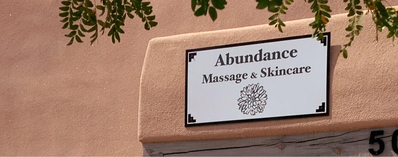 Sign for Abundance Massage & Skincare business, mounted on a peach-colored wall, with green leaves hanging above.