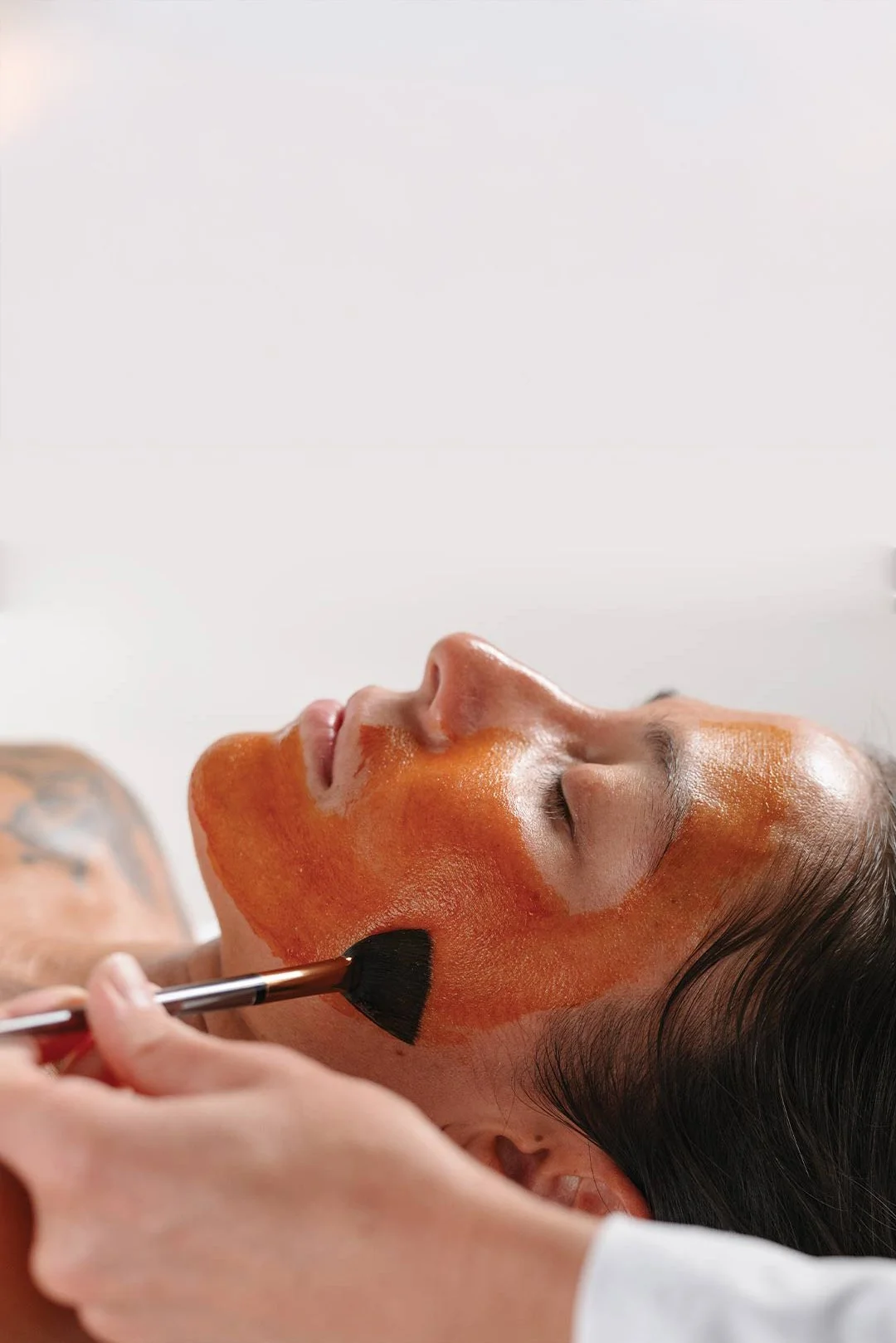 A woman receiving a facial treatment with a mask applied to her face by a skincare professional using a brush.