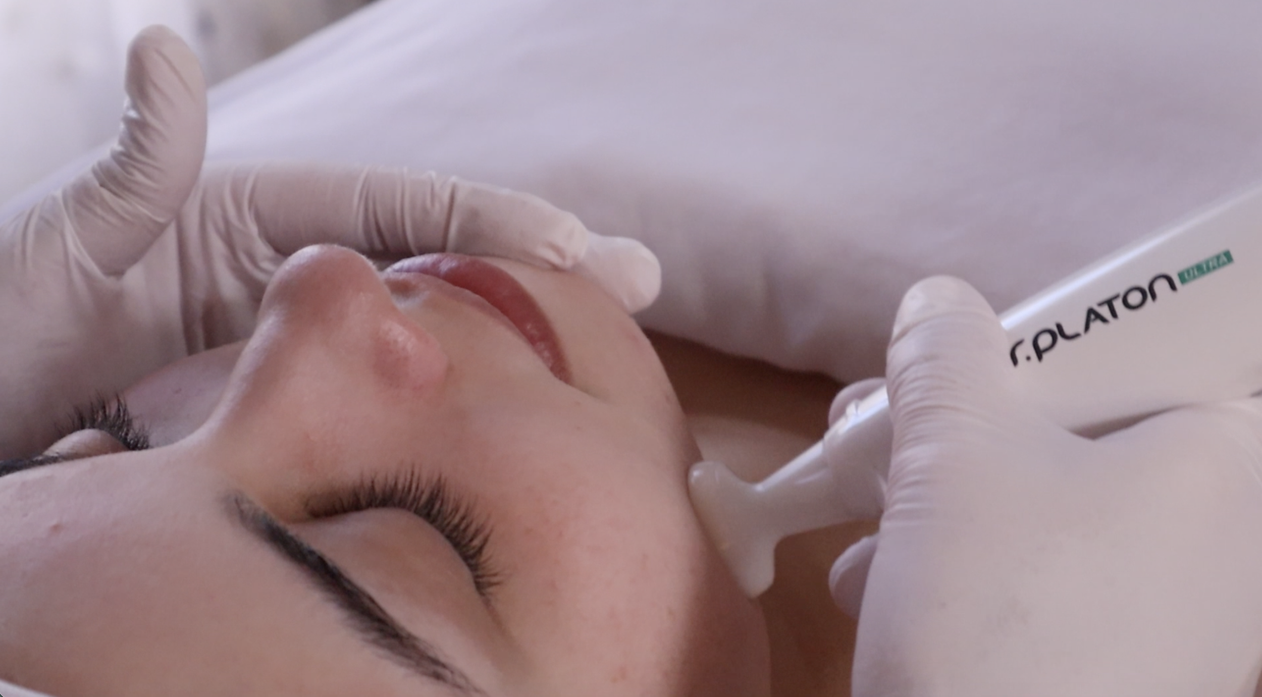 A person lying down with their eyes closed, receiving a cosmetic or medical treatment involving a device labeled r.PLATON, held by gloved hands.