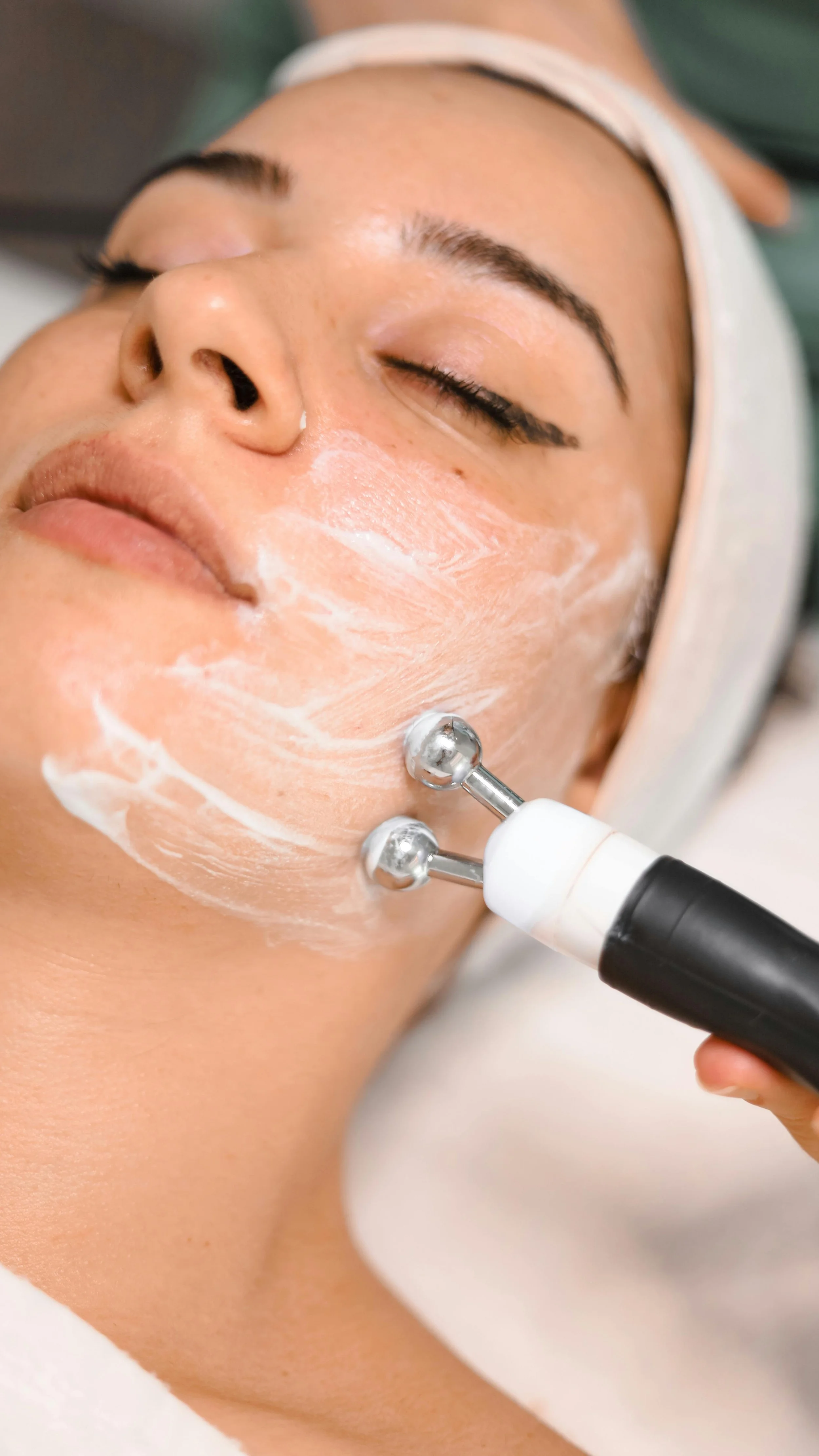 A woman lying down with her eyes closed gets a facial treatment using a handheld device with metal rollers on her cheek, with a white cream applied on her face.
