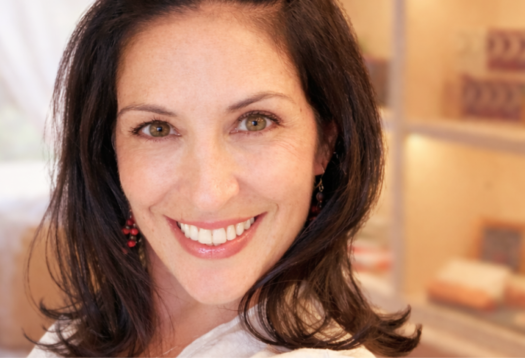 Close-up of a smiling woman with brown hair, wearing earrings and a white top, indoors with blurred shelves in the background.