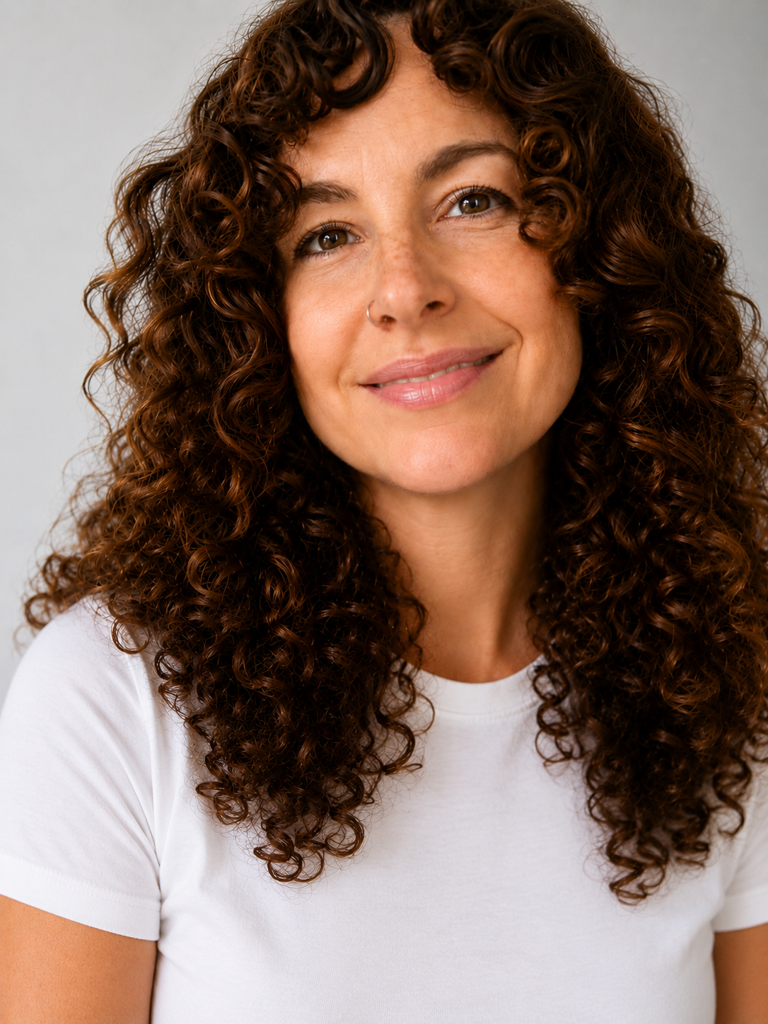 Close-up of a smiling woman with brown hair, wearing earrings and a white top, indoors with blurred shelves in the background.