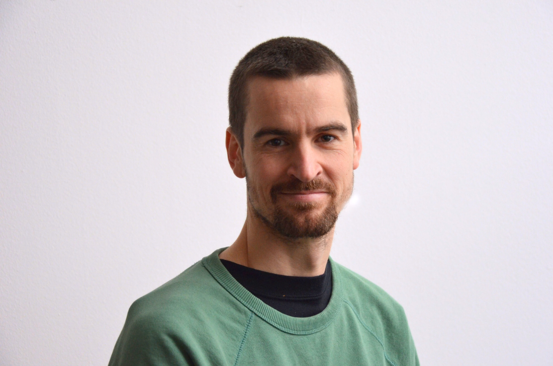 Ein Mensch mit dunklem, kurzgeschnittenem Haar und Bart, trägt ein grünes Sweatshirt, steht vor einer weißen Wand und lächelt leicht.