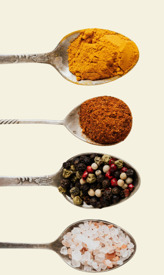 Four metal spoons arranged vertically containing different spices and salts: turmeric powder in the top spoon, ground cinnamon in the second, mixed peppercorns in the third, and Himalayan pink salt in the bottom spoon.