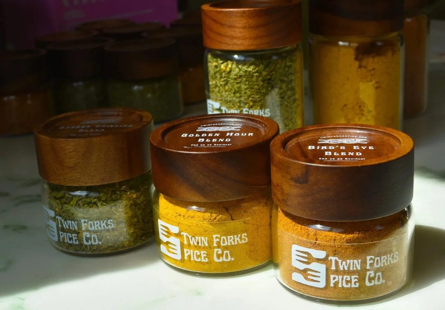 Jars of spices from Twin Forks Pice Co., labeled 'Golden Hour Blend', 'Bird's Eye Blend', and other spice blends, with some jars having wooden lids and others with glass lids, on a white surface.