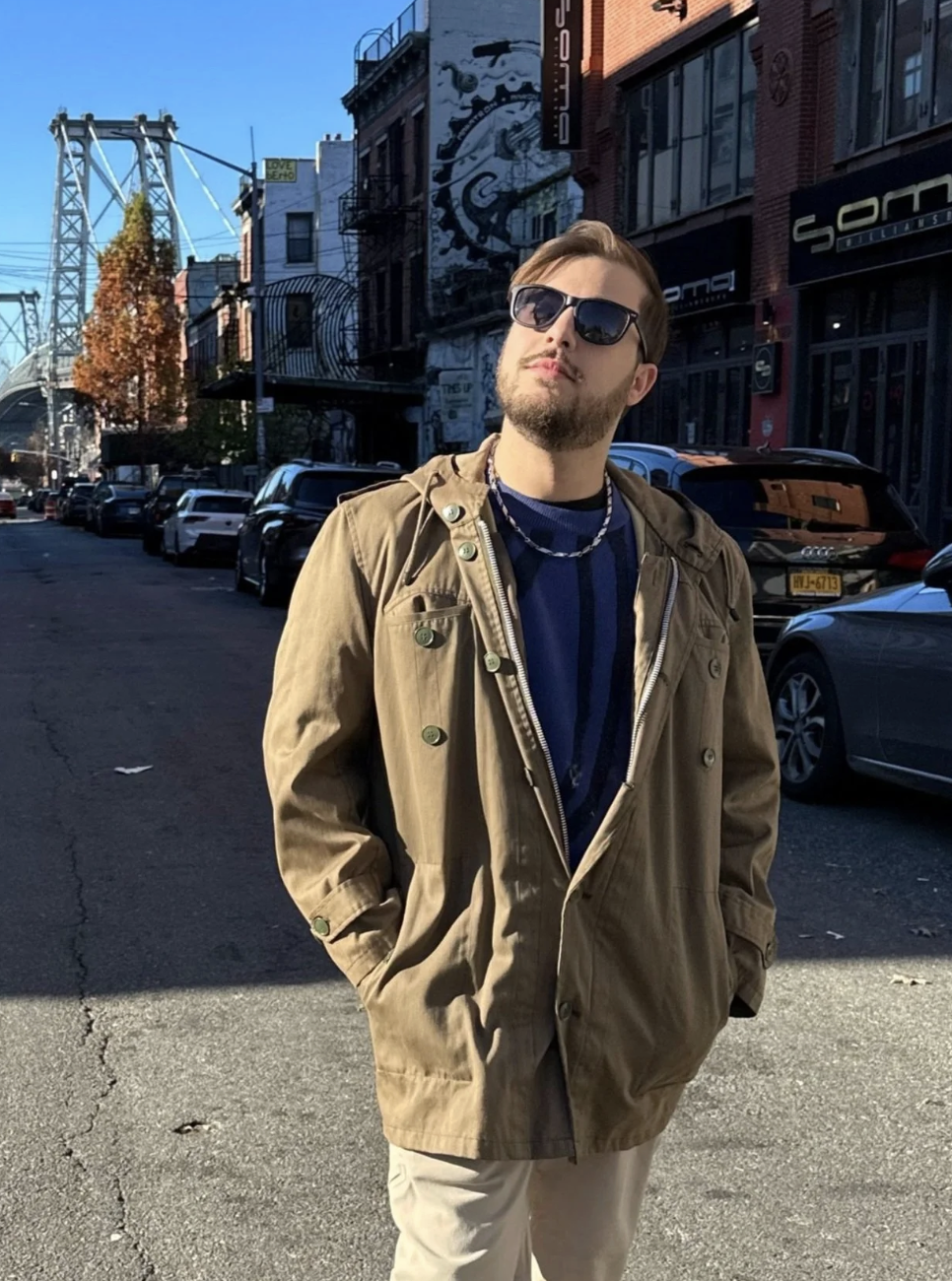 A young man with sunglasses and a beard standing on a city street with old buildings, parked cars, and a bridge in the background. He is wearing a tan jacket and a navy blue shirt.