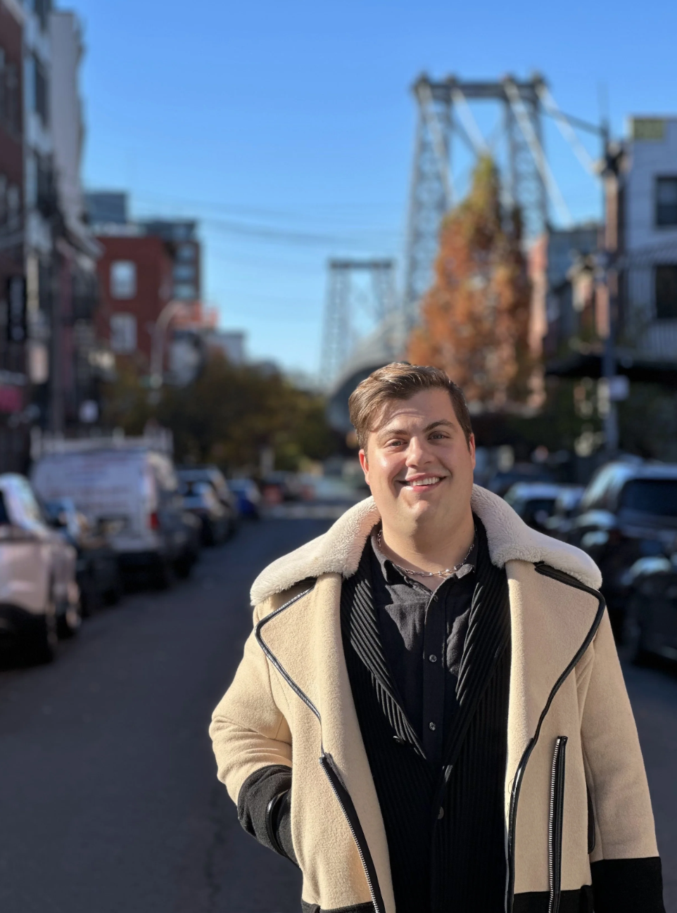 A smiling young man with short brown hair stands on a city street with parked cars and buildings in the background, including a bridge or tower structure and trees with autumn leaves, under a clear blue sky.