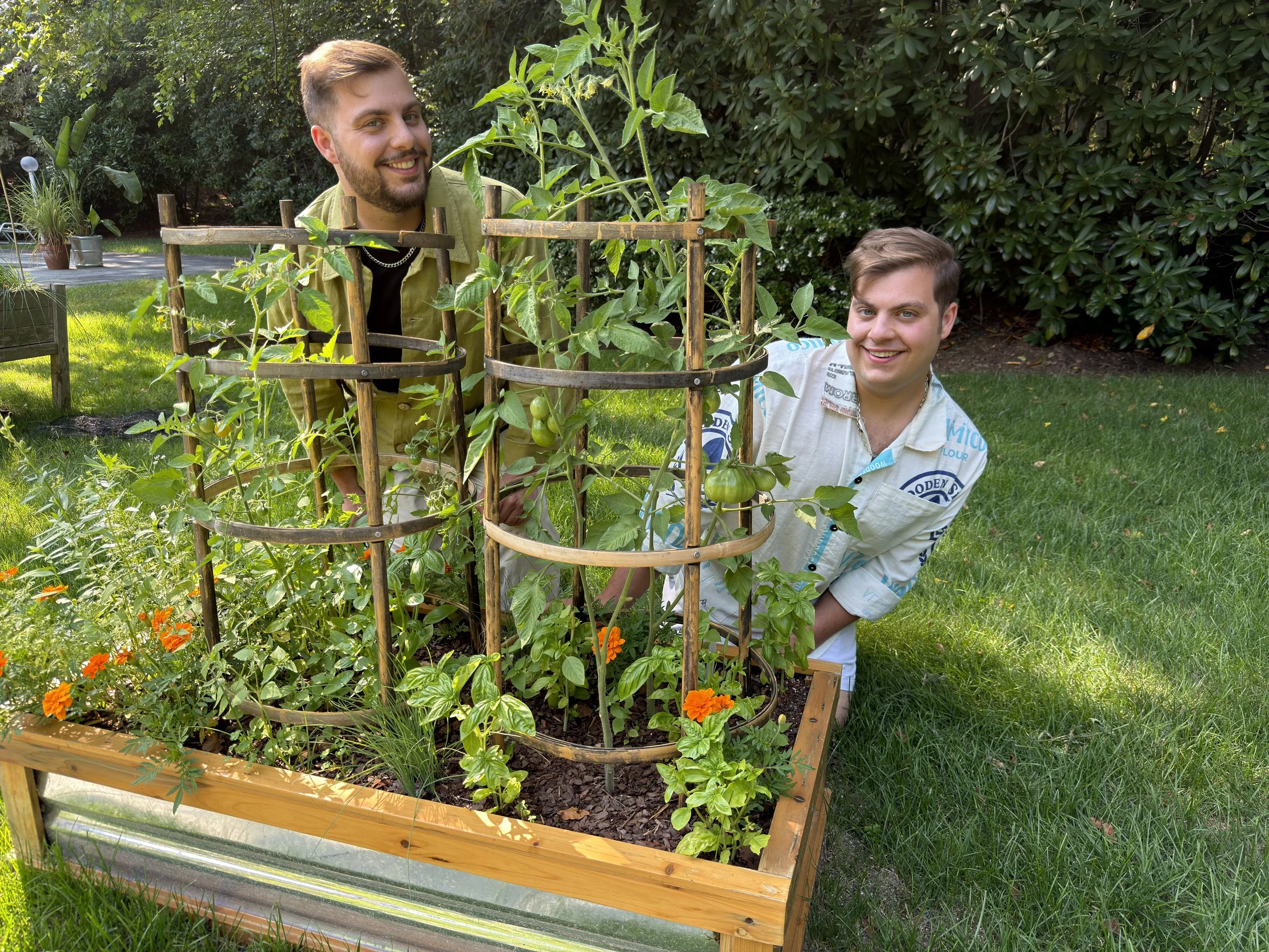 Two men pose behind a raised garden bed filled with tomato plants, basil, marigolds, and other vegetables.