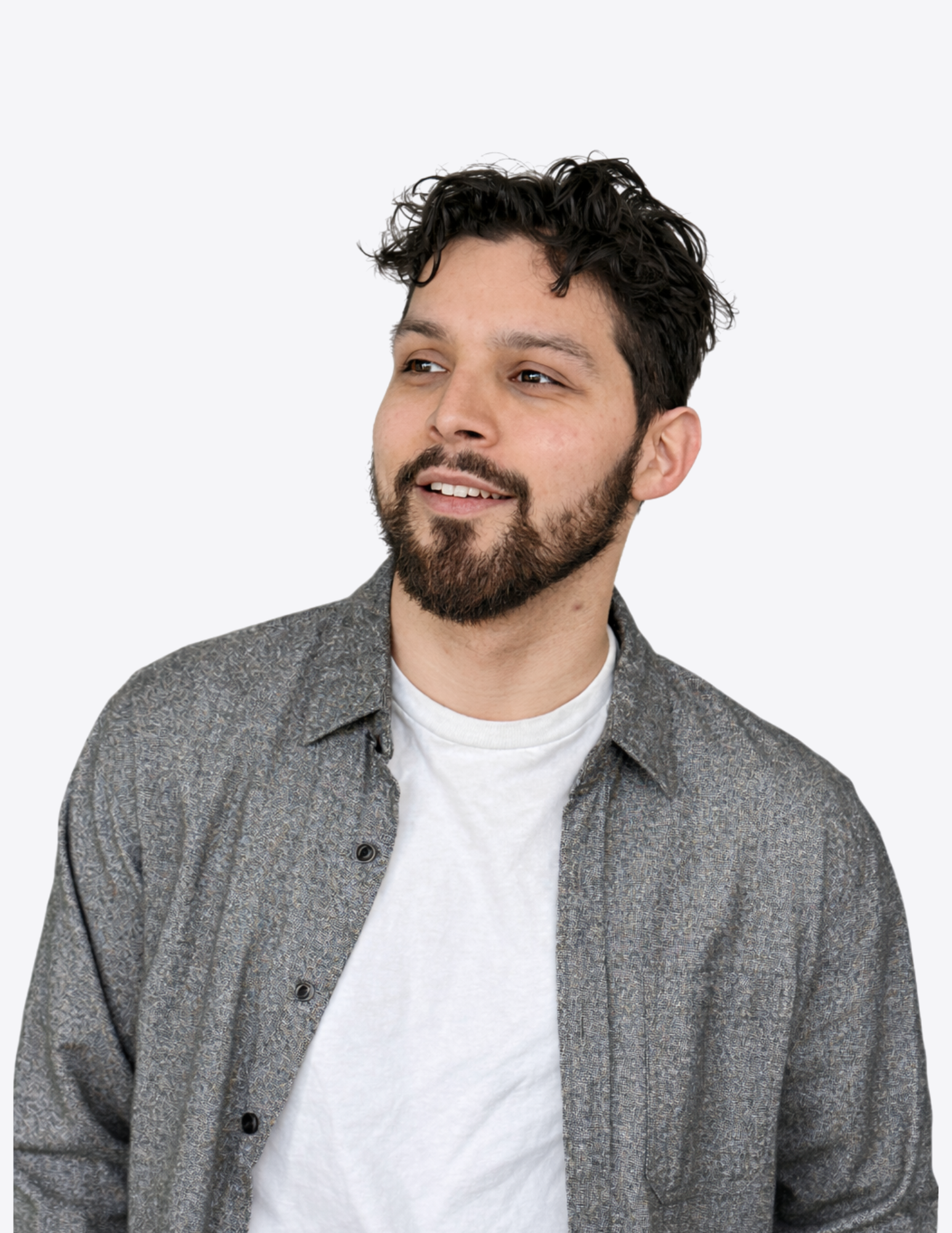 A young man with dark curly hair, a beard, and light skin, wearing a white T-shirt and a gray patterned button-up shirt, looking slightly to the side and smiling.