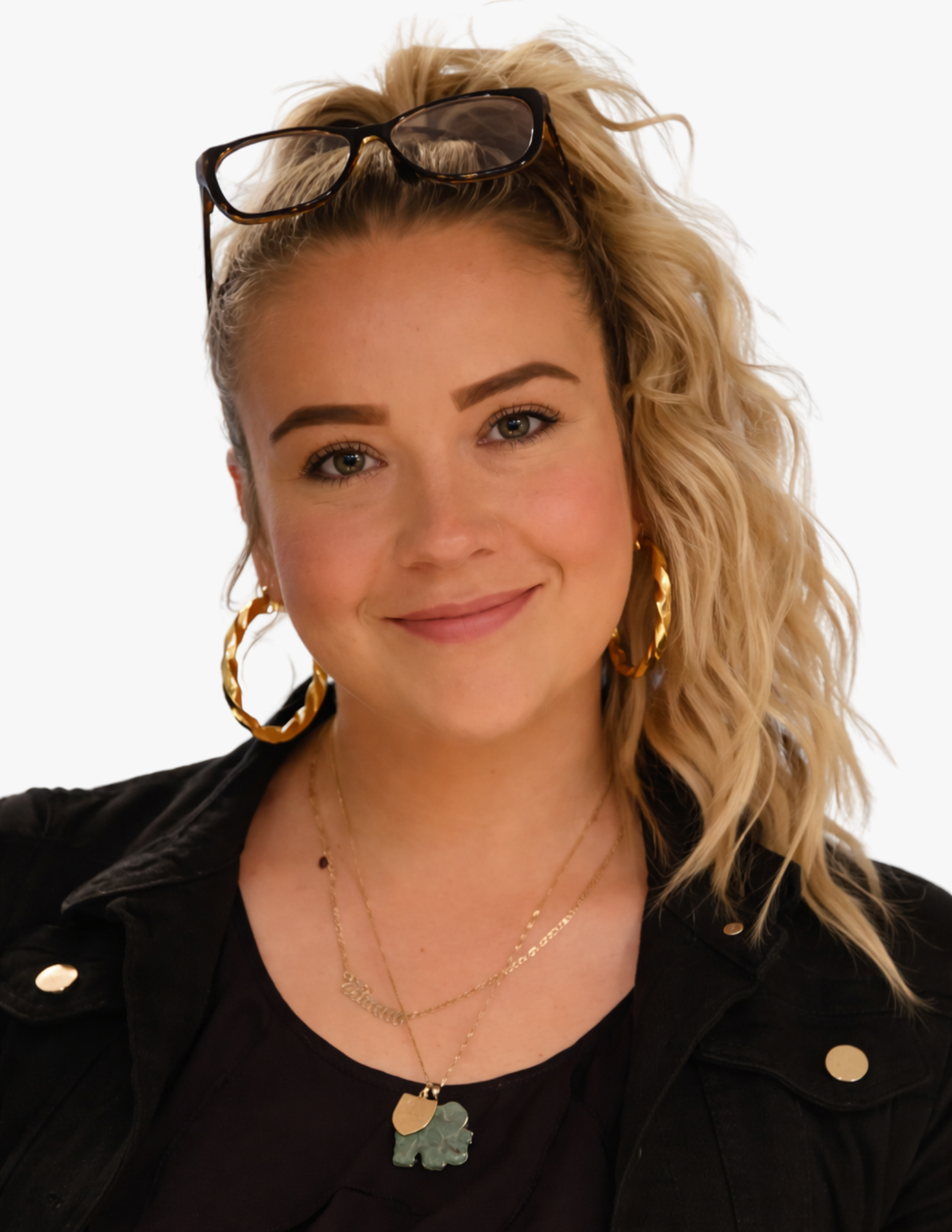 A young woman with blonde, curly hair styled in a half-up, half-down look, wearing glasses on her head, large gold hoop earrings, layered gold necklaces, and a black jacket with gold buttons, smiling at the camera.