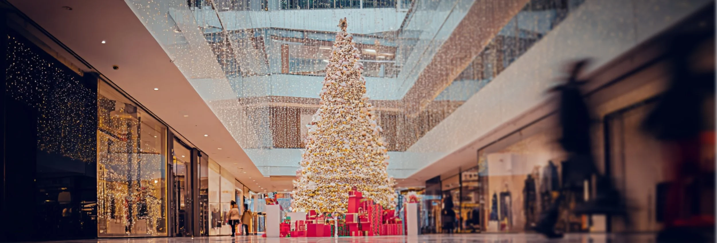 Large Christmas tree decorated with lights and ornaments, surrounded by wrapped presents, in a shopping mall with glass ceiling and storefronts.