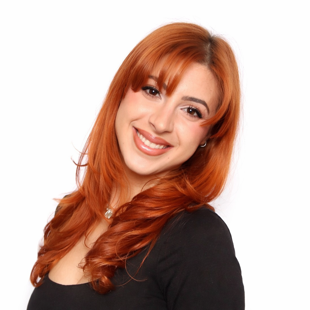 A young woman with long, curly red hair and light skin smiling at the camera, wearing a black top and small hoop earrings, against a plain white background.