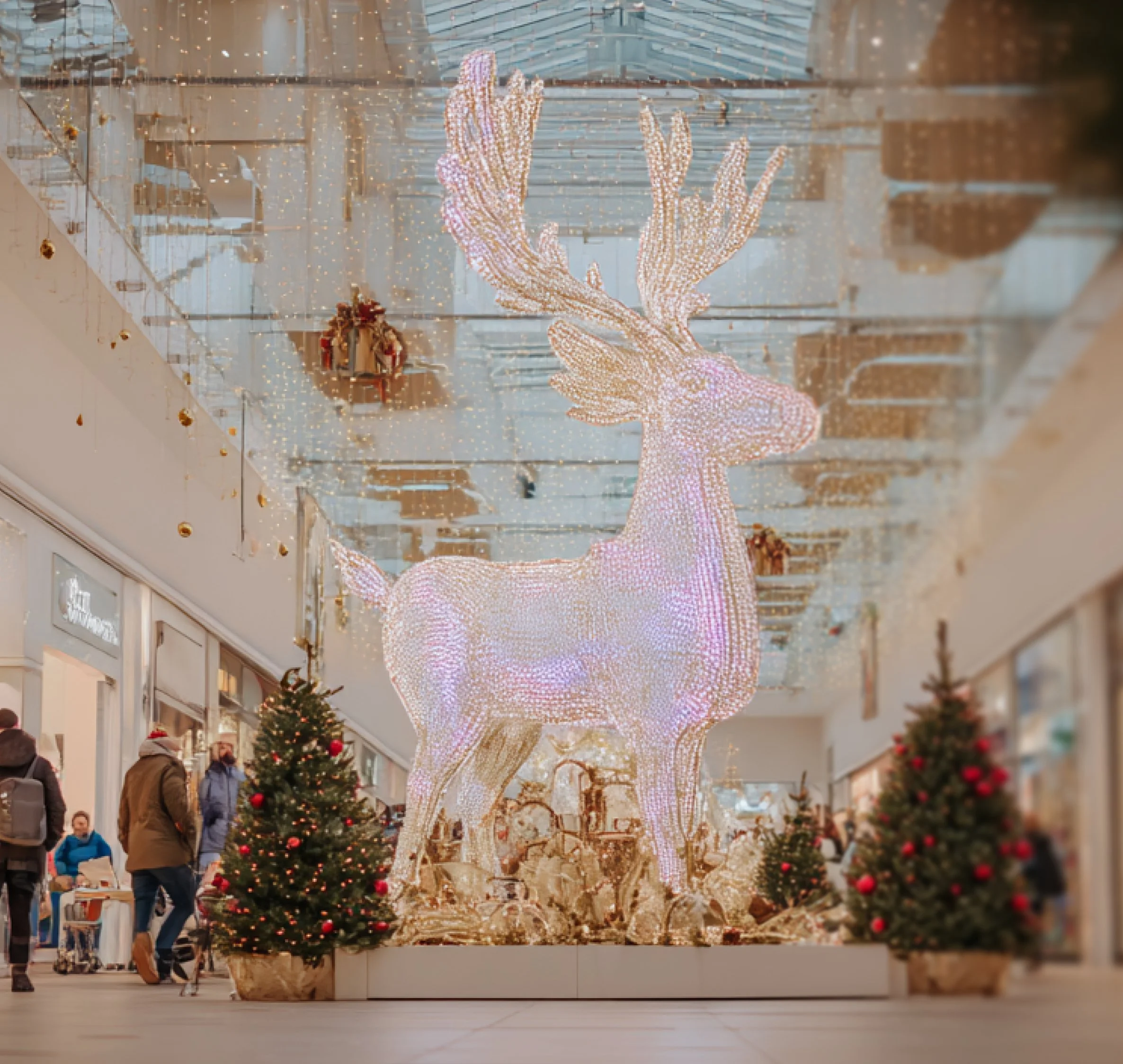 Illuminated decorative reindeer with antlers in a shopping mall, surrounded by Christmas trees and festive decorations.