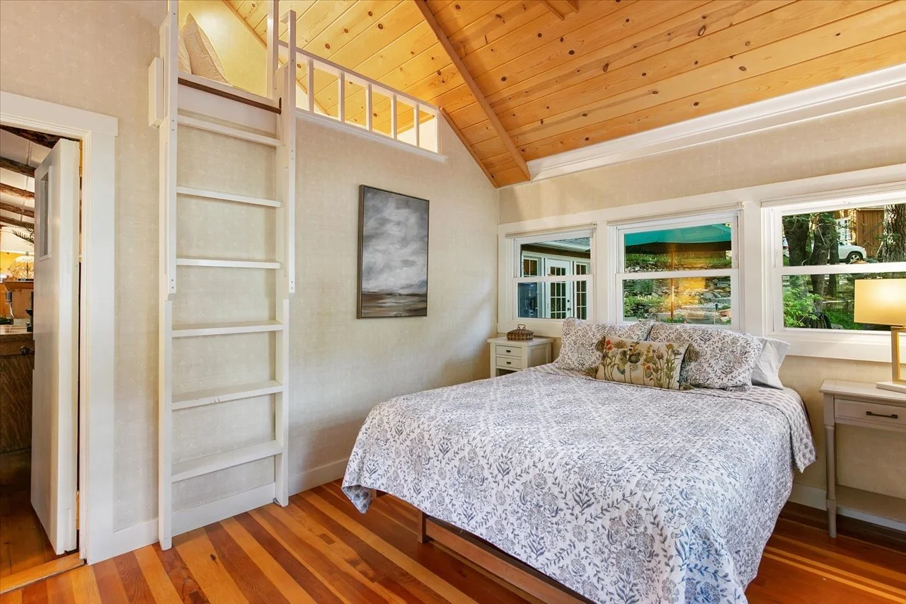 A cozy bedroom with a wooden vaulted ceiling, large windows, a bed with floral bedding, white nightstands, and a small loft space accessible by a white ladder.