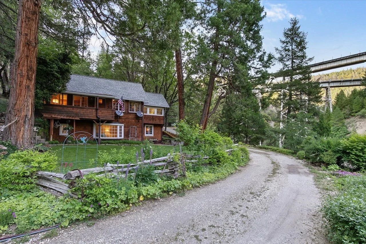 A rustic wooden house with a gray roof and balcony, surrounded by lush green trees, beside a gravel road with a wooden fence and garden plants, with a bridge and forested hills in the background.