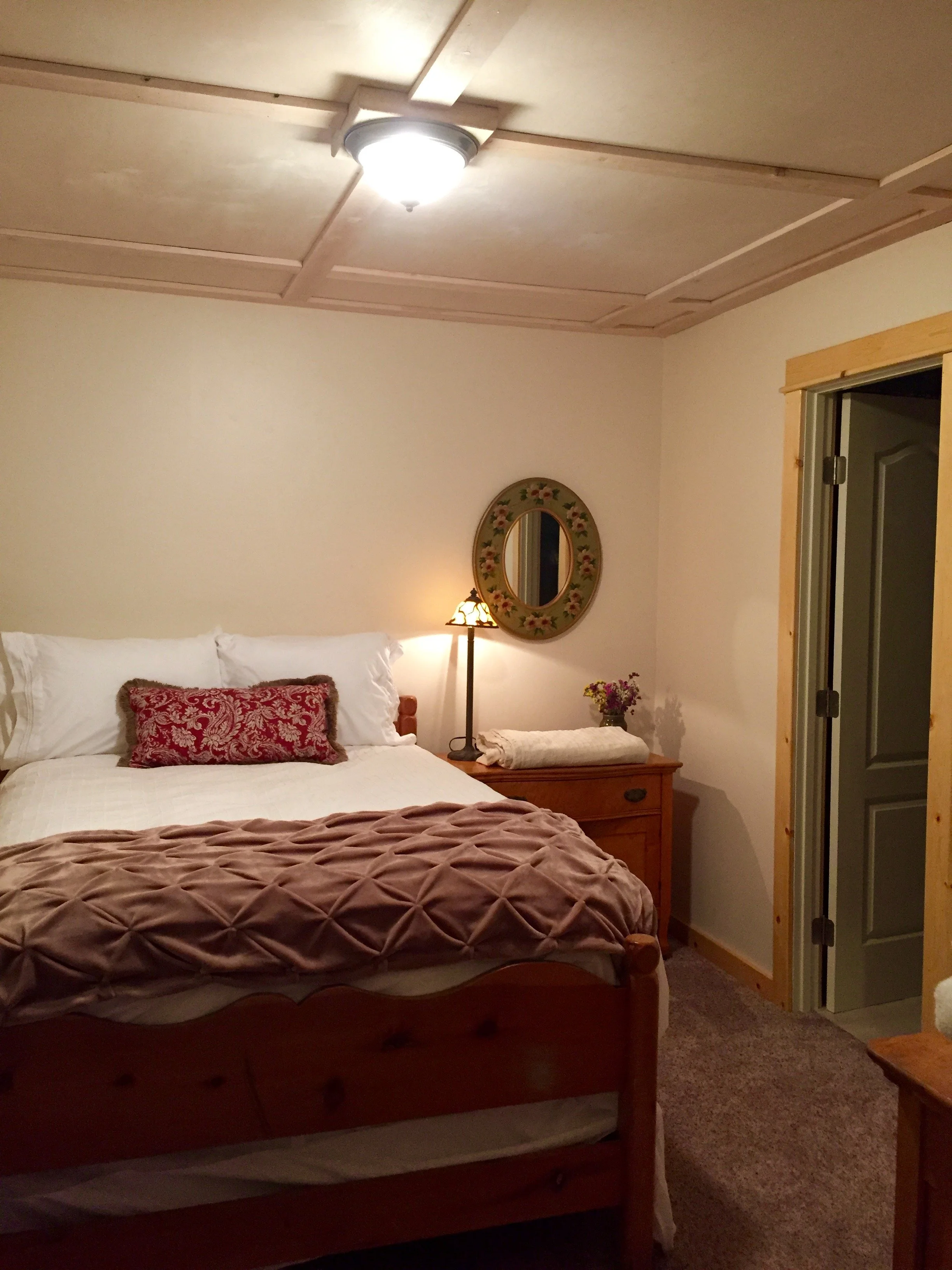 Cozy bedroom with a wooden bed, white bedding, pink plush blanket, and decorative pillow. A nightstand with a lamp, flowers, and towel. Mirror on the wall and door to a closet.