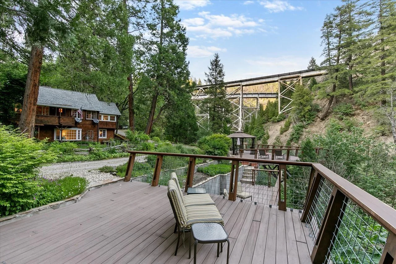 View from a wooden deck overlooking a lush green yard with a cabin-style house on the left, tall trees, and a railway bridge in the background, under a partly cloudy sky.