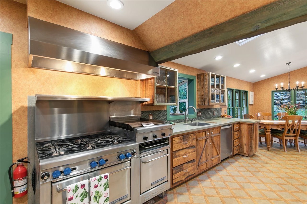 Kitchen with stainless steel stove and oven, wooden cabinets, green window frames, and a dining area with wooden chairs and a chandelier.