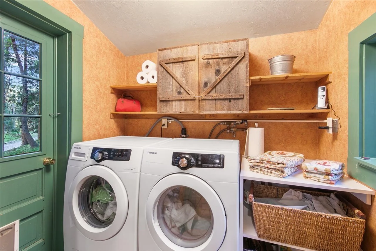 A laundry room with a front-loading washing machine and dryer, two green-framed windows, and wooden shelves holding paper towels, a red bag, a metal bucket, a cordless phone, and folded linens, with a wicker basket underneath the linens.
