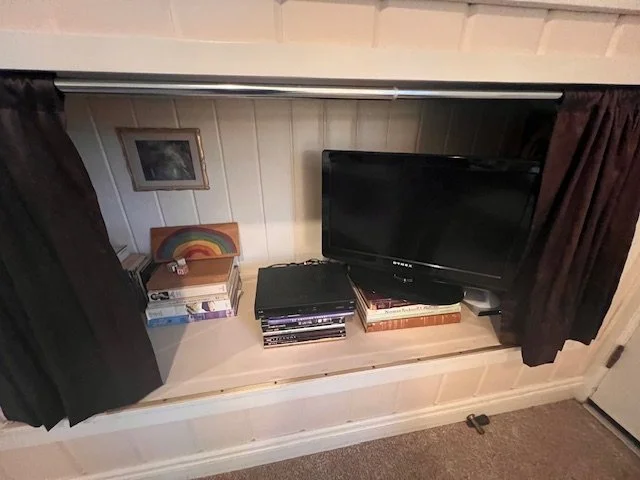 A small TV on a white shelf with a stack of DVDs underneath. To the left of the TV are stacked books and a small rainbow-themed decor piece. There is a picture hanging on the wall behind the shelf, and the area is framed by dark curtains on either si