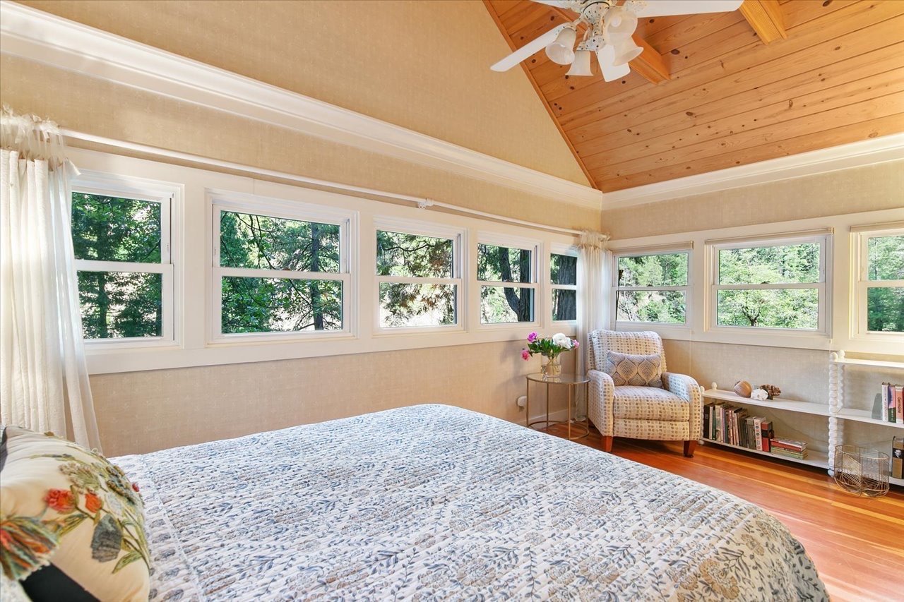 A bedroom with a bed covered in a patterned quilt, a floral pillow, and a wooden ceiling with a ceiling fan. There is a window beside the bed, a chair, a small table with flowers, and a white bookshelf with books.