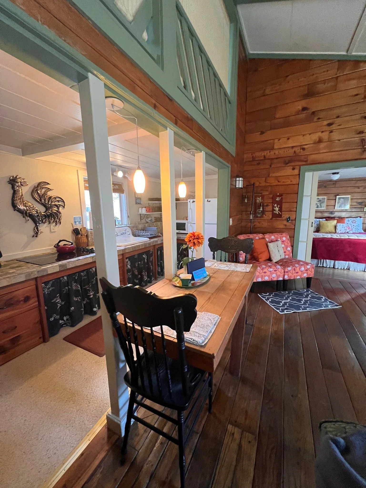 Interior view of a cozy home with a dining area, kitchen, and bedroom feature visible. The dining table has a flower centerpiece and placed settings, with black chairs. The walls are wood-paneled, creating rustic charm. There is a doorway leading to 