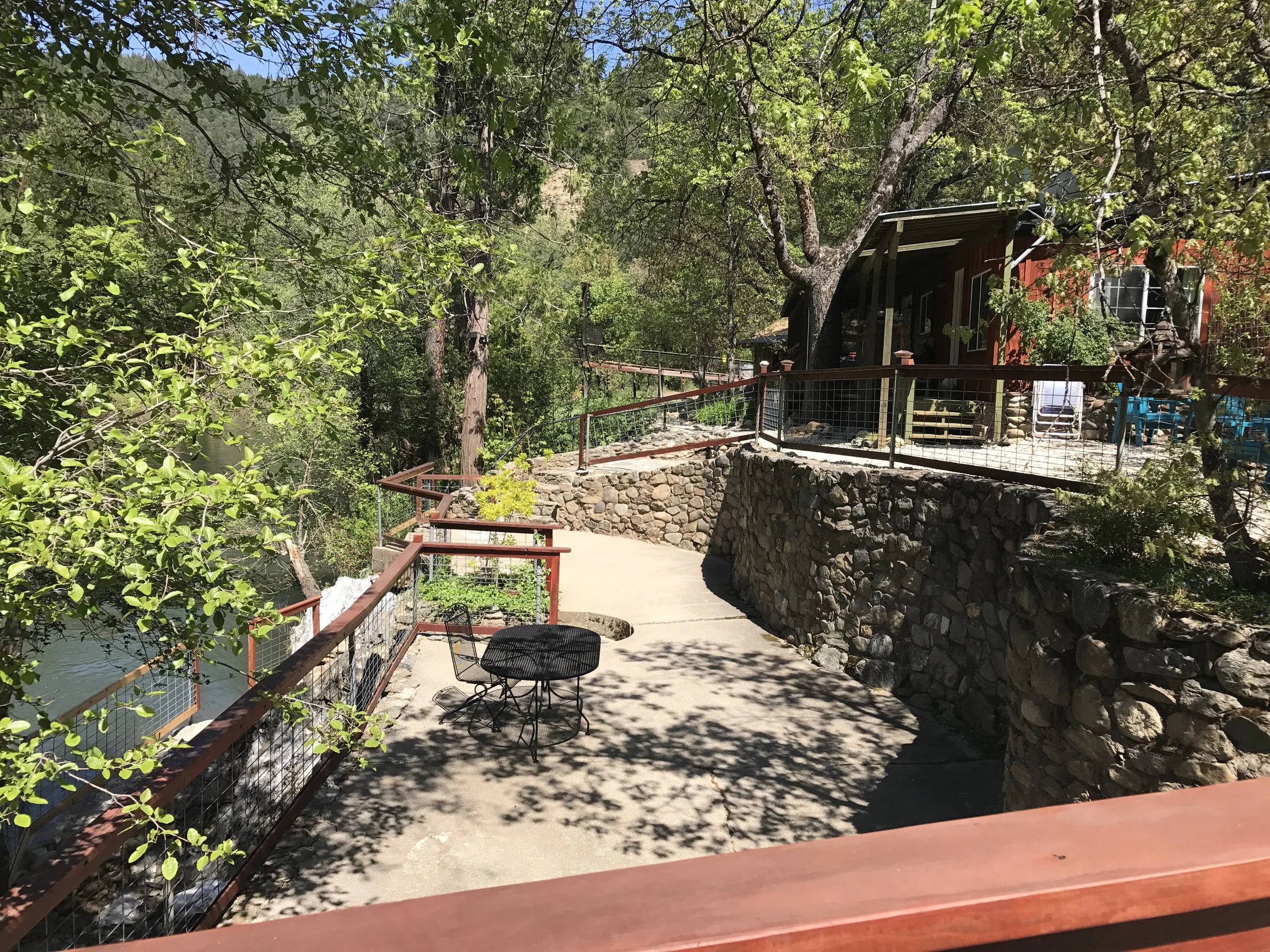 A cabin with a red exterior and a porch, situated among green trees. There is a concrete pathway bordered by a stone wall, with a small round table and a chair. Shadows of leaves are cast on the pathway, and the area appears peaceful and scenic.