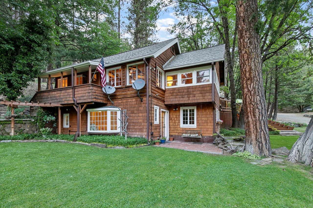 A two-story wooden house in a lush green yard surrounded by trees, with satellite dishes attached to the upper level and an American flag on the porch.