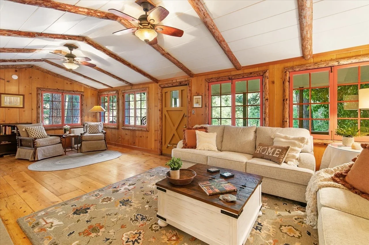 Living room with wooden walls and ceiling, two ceiling fans, large windows, beige sofas, armchairs, a coffee table with plants and books, and a patterned area rug.