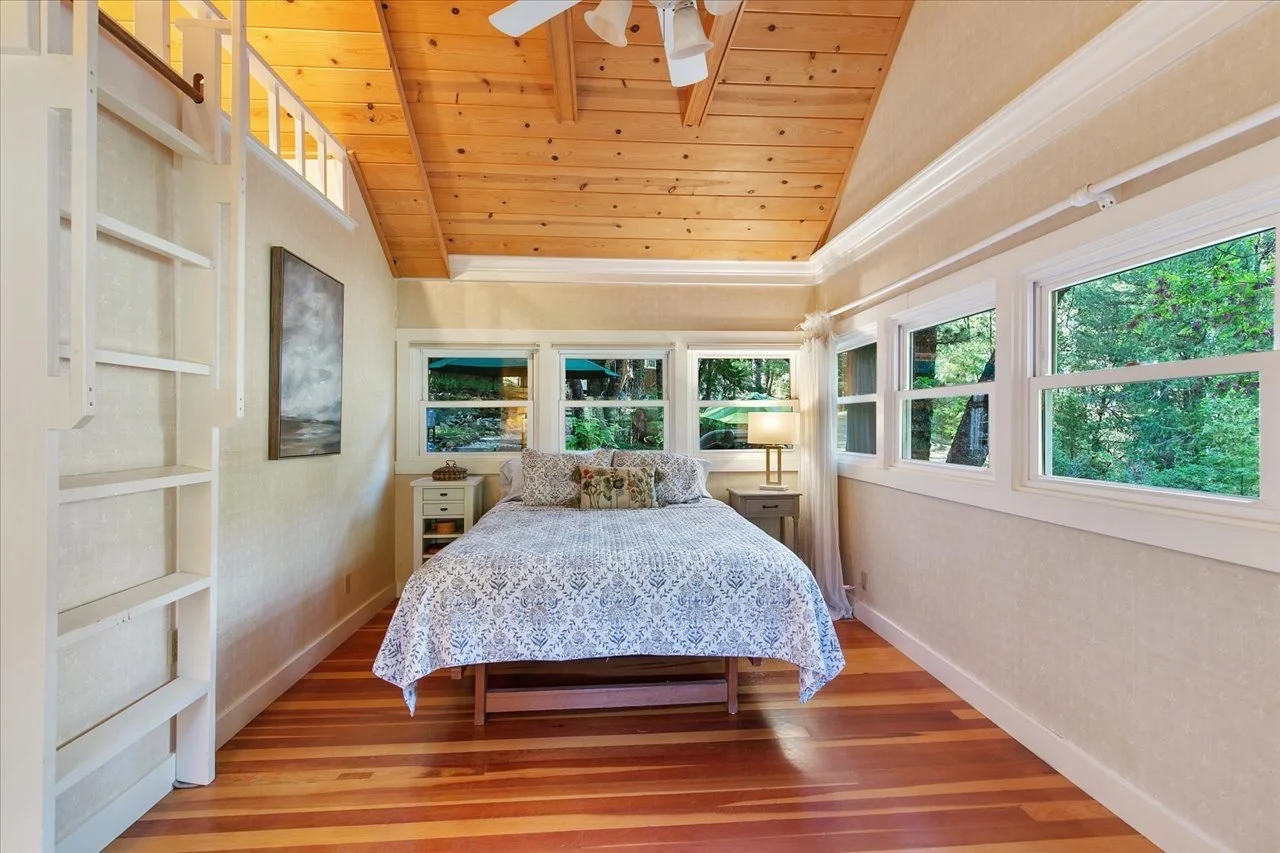 A cozy bedroom with a wooden ceiling, multiple windows with views of greenery, a bed with a patterned quilt, two side tables with lamps, and a painting on the wall.