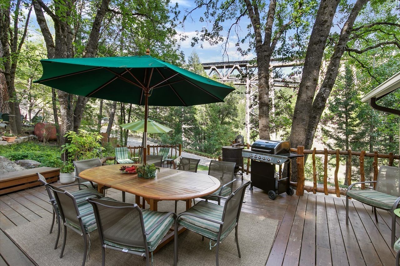 Outdoor wooden deck with a dining table, chairs, a large green umbrella, and a barbecue grill, surrounded by trees and greenery.