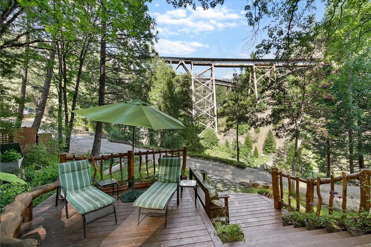 A wooden deck with two green striped lounge chairs and a green patio umbrella, overlooking a forested landscape and a large railway bridge in the background.