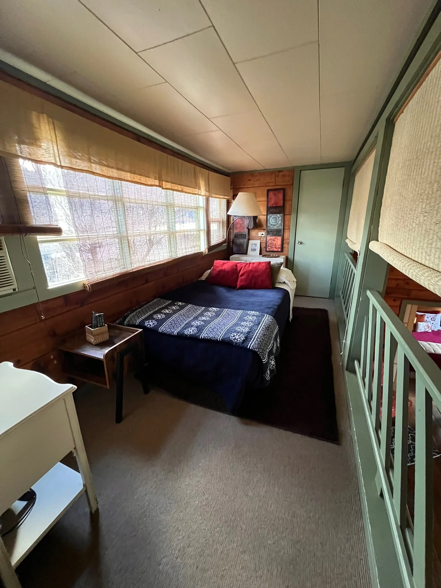 Small bedroom with single bed, nightstand, lamp, window with blinds, wooden wall, and partial view of a loft area.