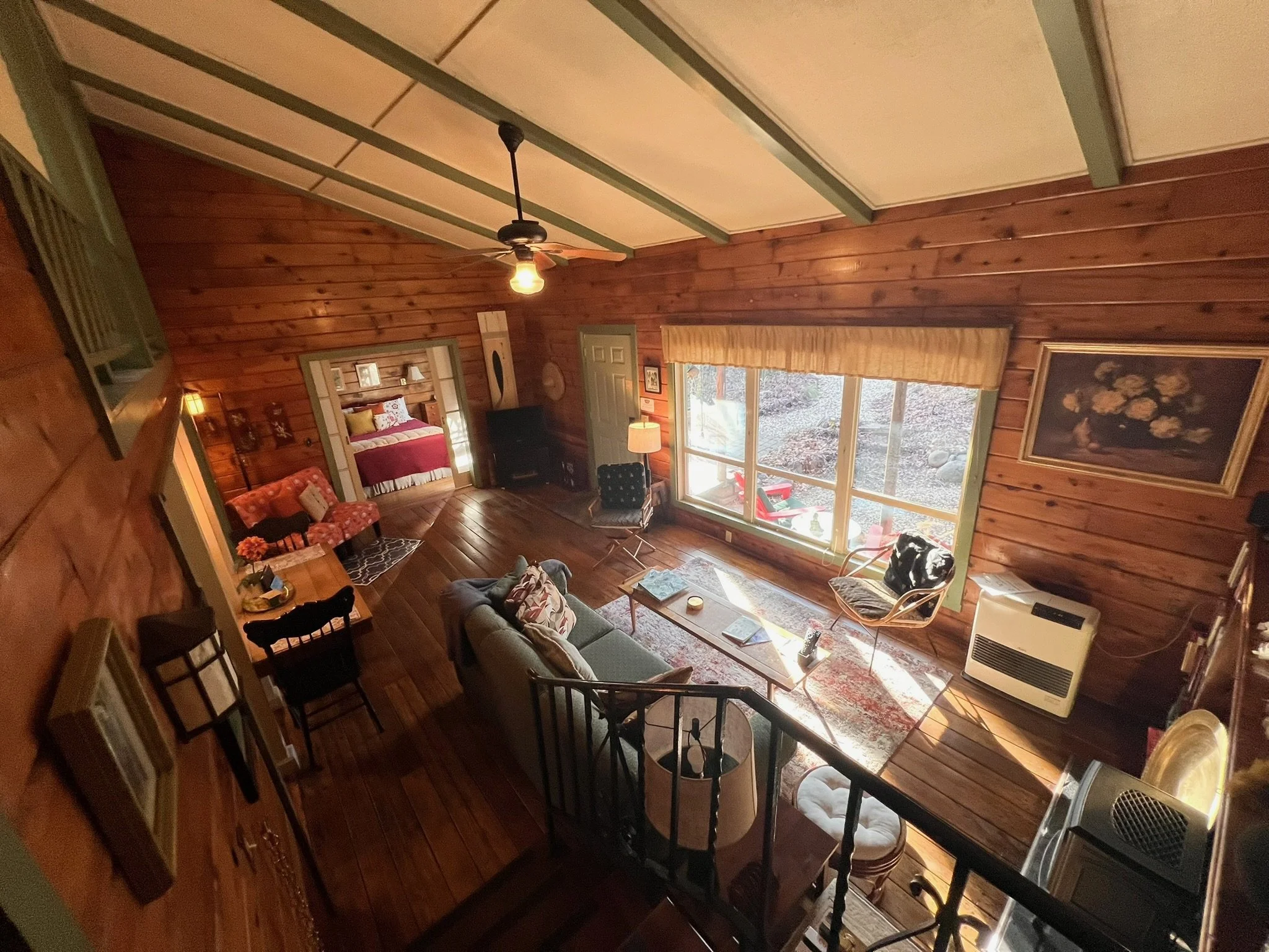 Inside a cozy, rustic living room with wood-paneled walls, large window, and various furniture including sofa, armchairs, and a dining table.