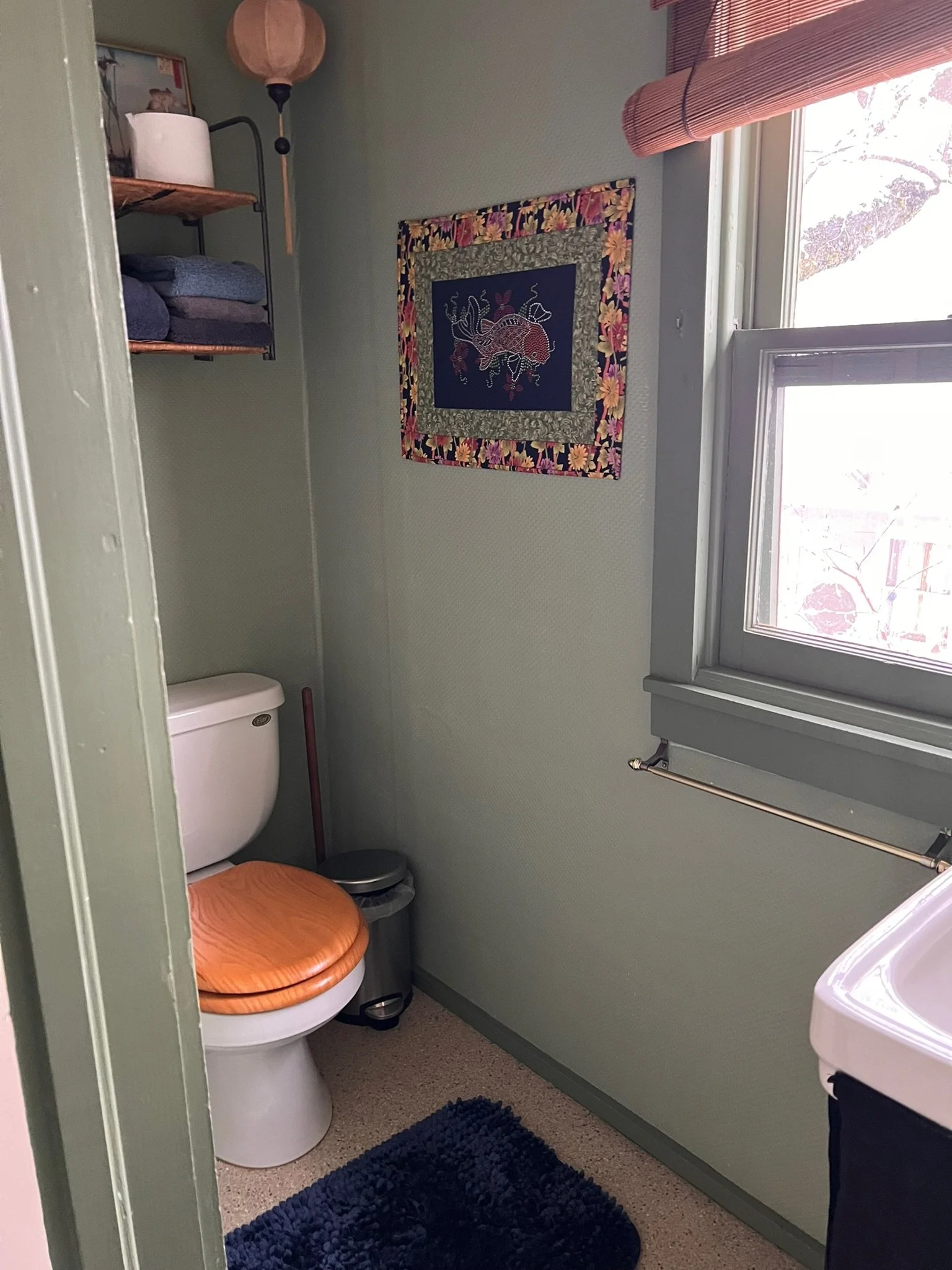 Small bathroom with green walls, a toilet with a wooden seat, a black rubber mat, a window with brown blinds, a wall shelf with folded towels, a floor lamp, and decorative wall art.