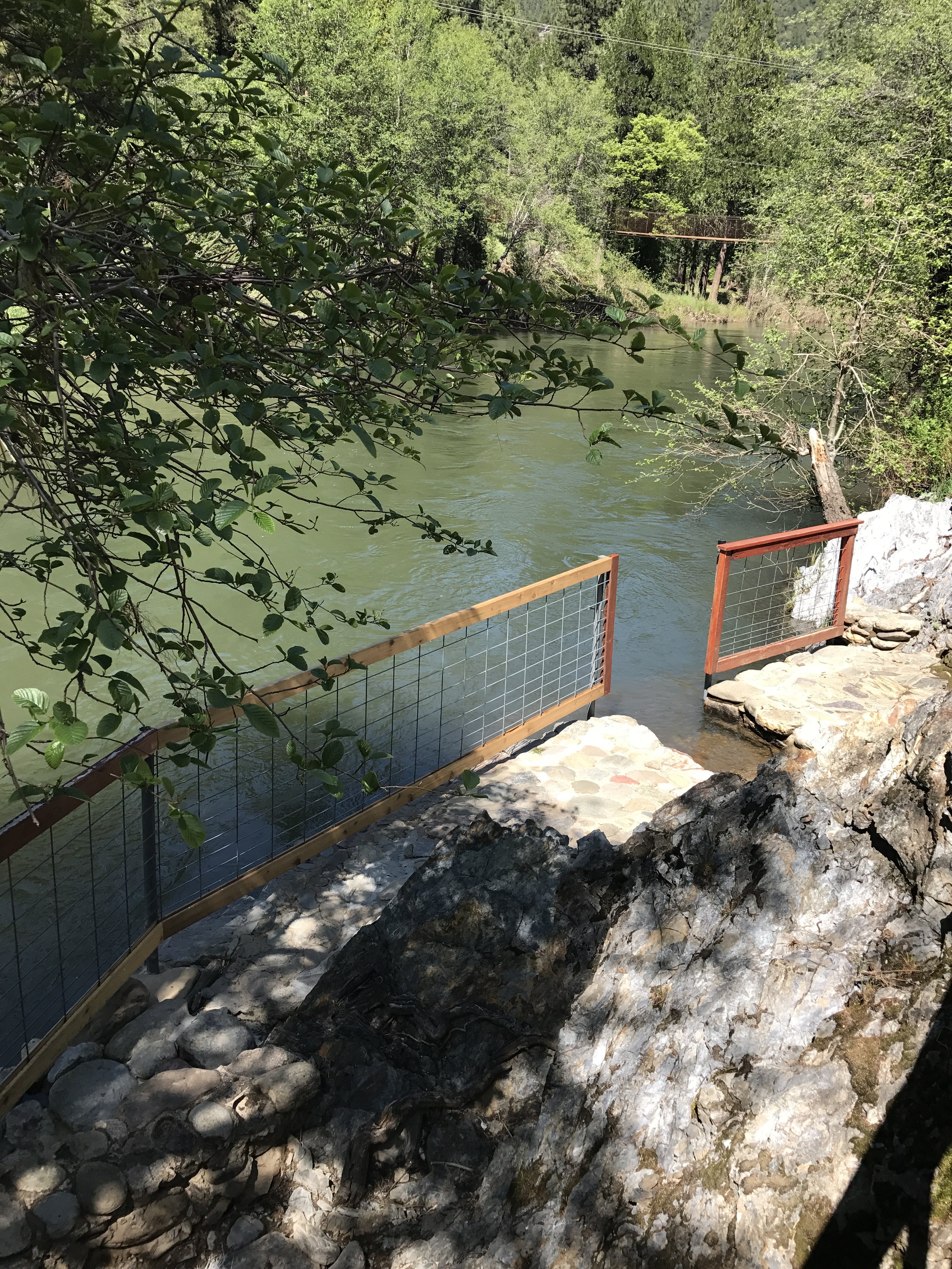 A river with surrounding green trees, a rocky shoreline, and a small wooden fence with wire mesh along the edge of the water.