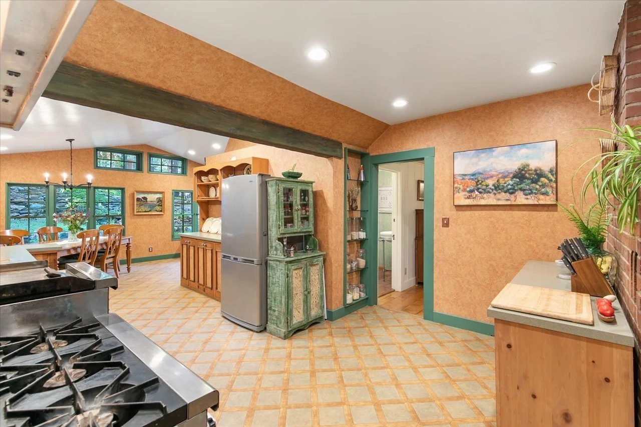A kitchen with orange textured walls, a tiled floor, and green trim around the doorway and windows. The kitchen includes a stainless steel stove, a blue-green wooden cabinet, a refrigerator, and a kitchen island. In the background, a dining area with