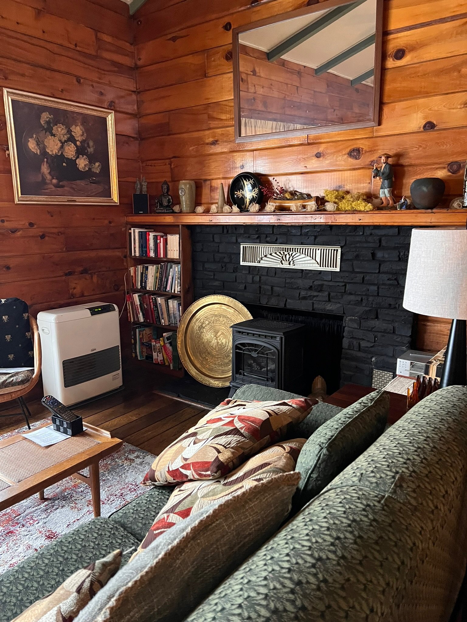 Living room with wooden walls, black brick fireplace, wooden shelves and furniture, and a framed floral painting. Items on the mantle include decorative objects and a small figurine. There's a bookshelf filled with books, a large golden tray, and a h
