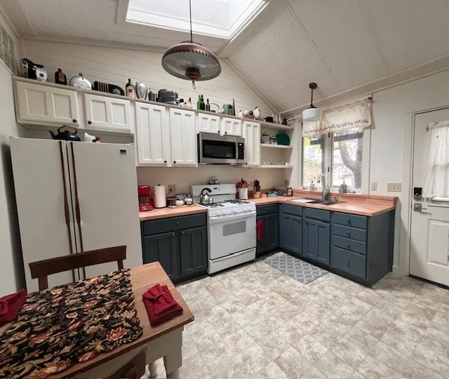 Kitchen with white upper cabinets, dark lower cabinets, a white refrigerator, and a white stove. There is a window above the sink, a skylight in the ceiling, and a wooden table with red napkins and a patterned runner.