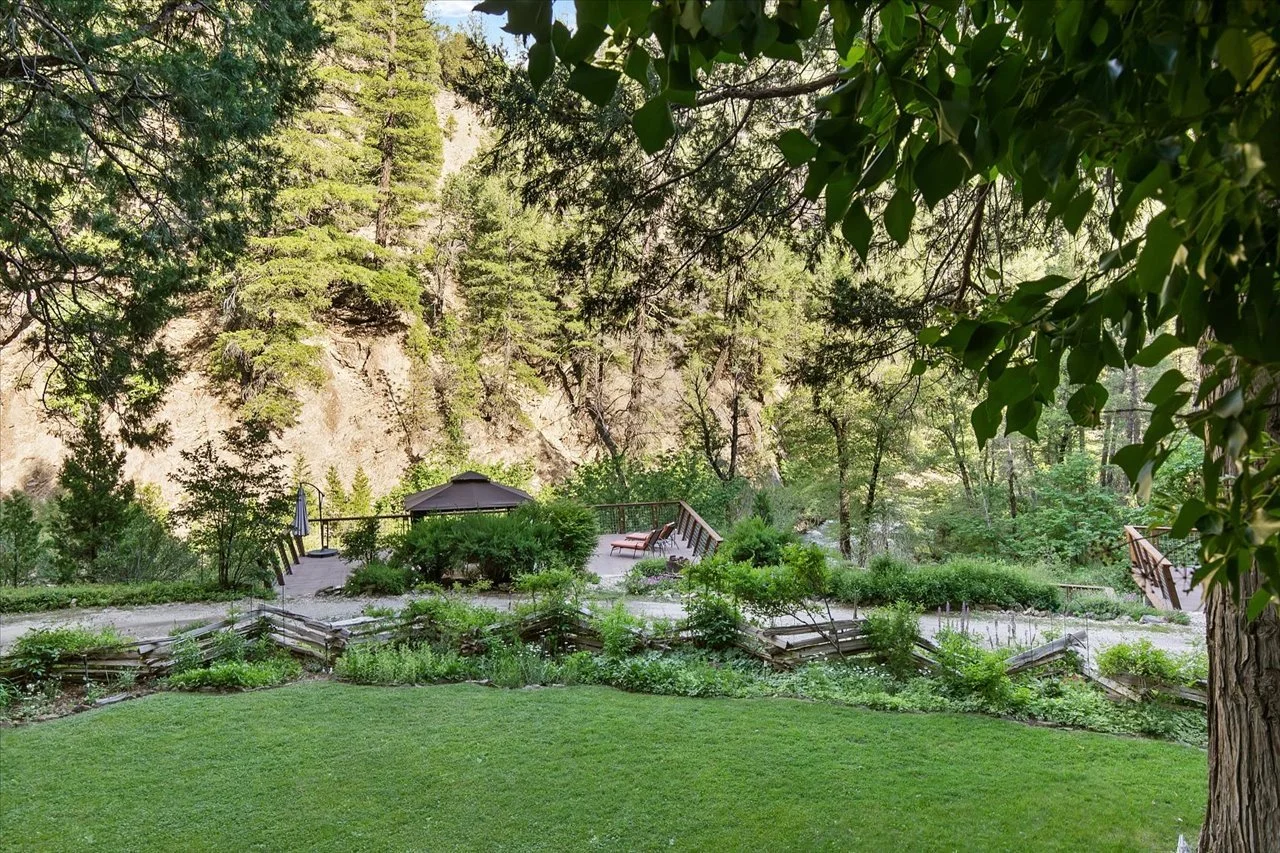 A lush green backyard with a grassy lawn and a garden area surrounded by trees and shrubs. There is a wooden deck with lounge chairs and a small patio umbrella, set against a backdrop of a rocky hillside covered with trees.