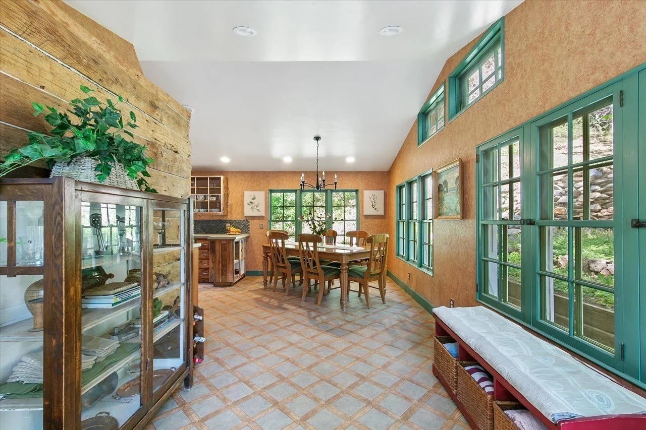 A dining room with large green-framed windows and a wooden dining table with six chairs. There is a chandelier above the table, and a wooden cabinet on the left. The walls are painted in a warm orange color, and the floor has a patterned tile design.